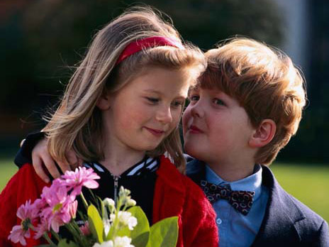 Color photo of a little boy whispering to a girl holding flowers
