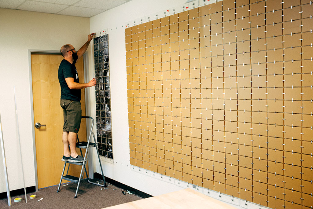 Person standing on a ladder assembling a printed photo mosaic, one photo at a time