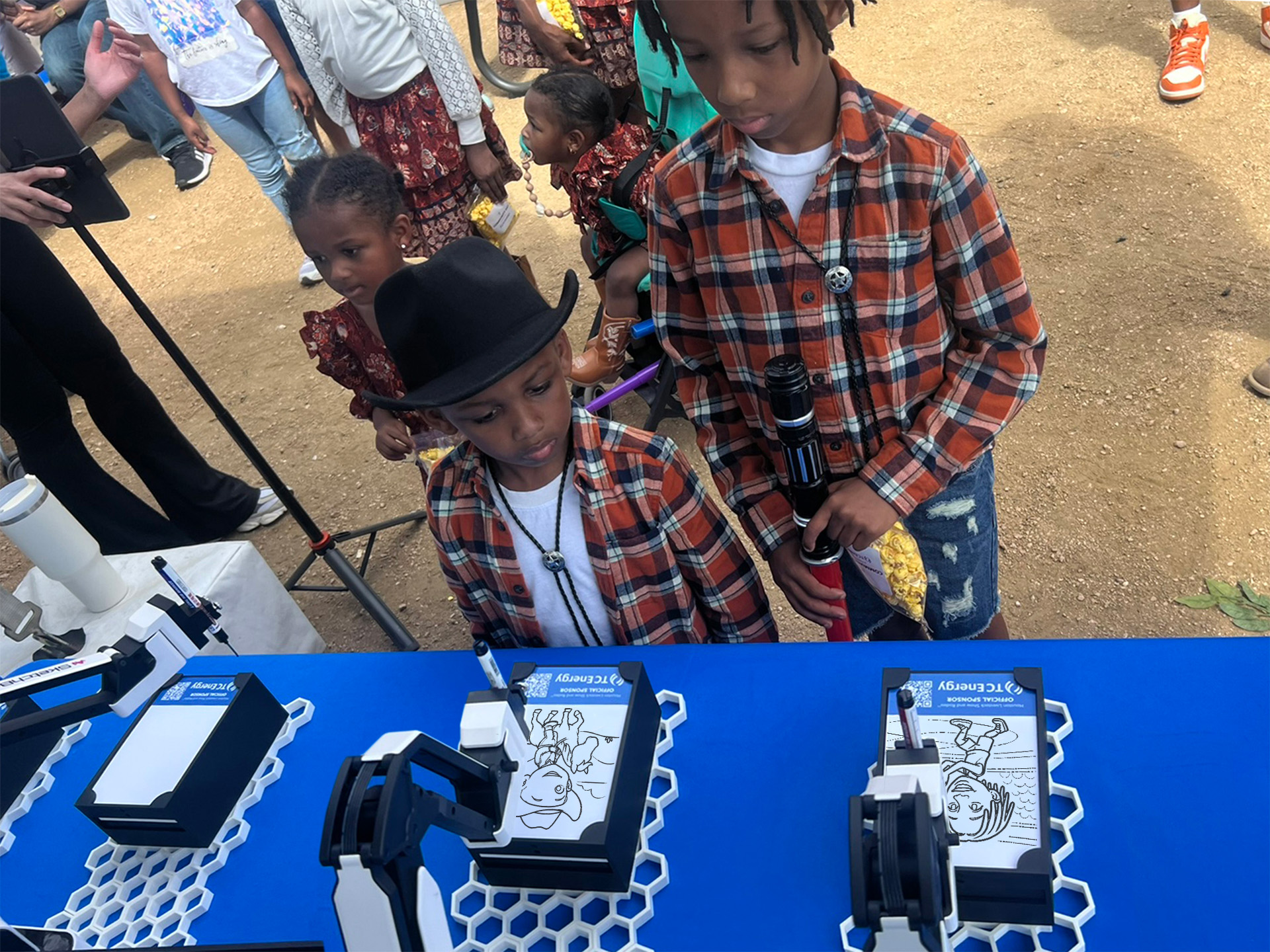 Two young boys dressed in cowboy hats watching the SketchBots draw their portraits